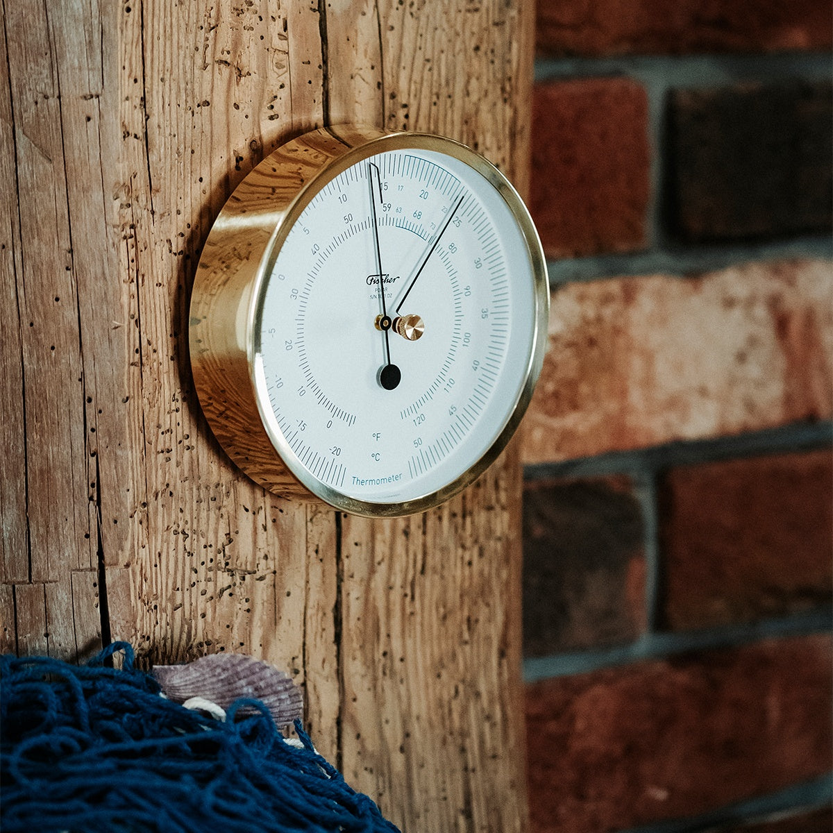 Brass analog wall thermometer with white dial on rustic wooden post, nautical decor