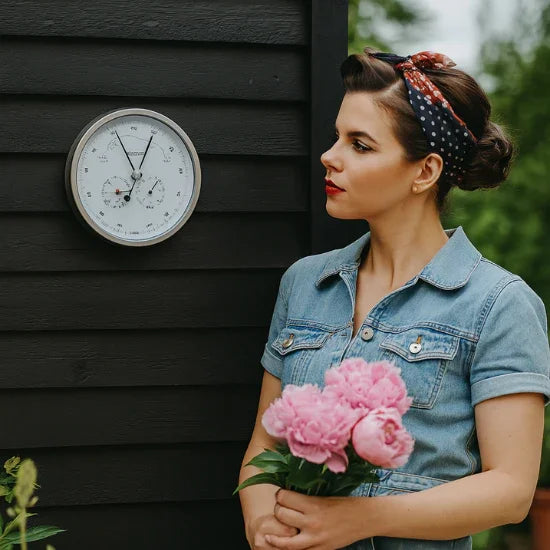Woman holding pink flowers standing by outdoor wall-mounted barometer and thermometer